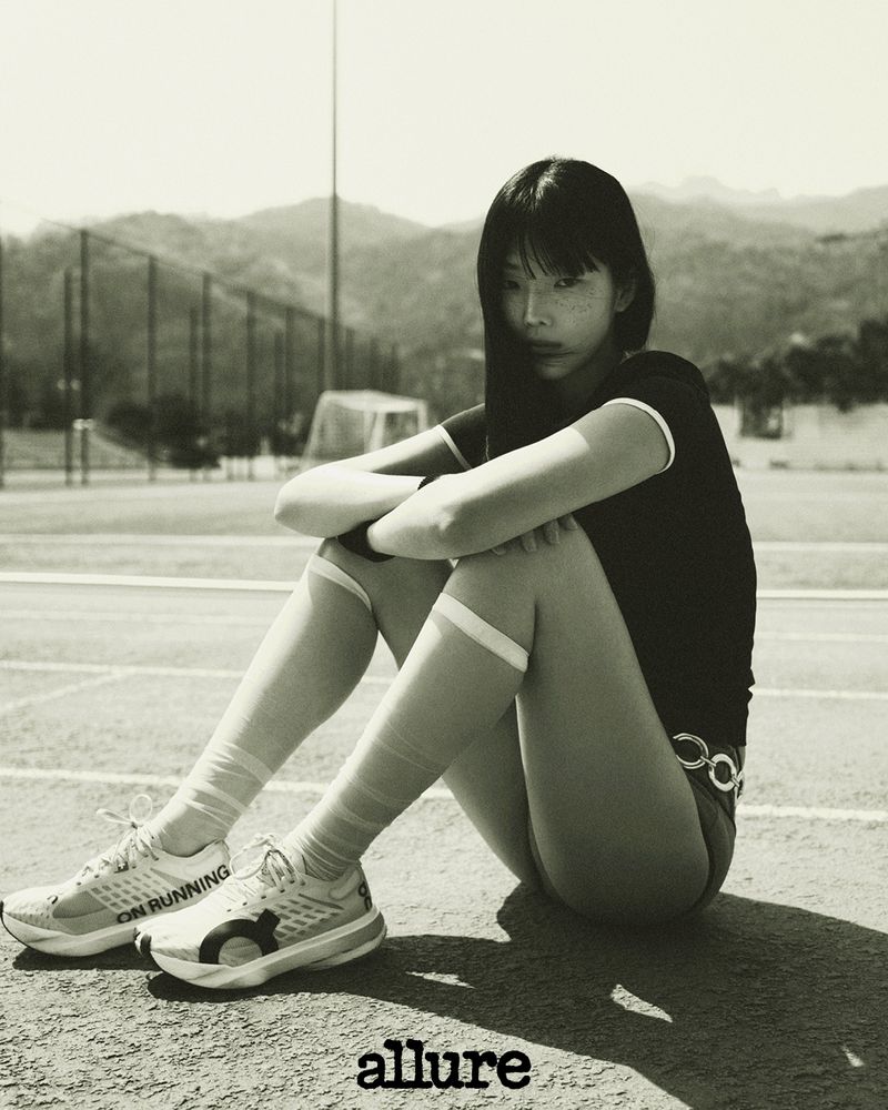 Athletic woman in sportswear sits on the track with running shoes in monochrome, mountains visible in the background under bright daylight.