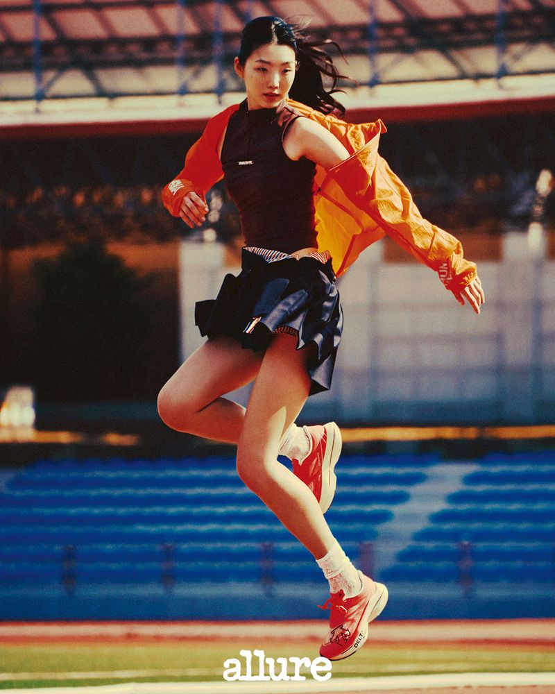 Athletic woman jumps mid-air on a running track, wearing a black top, navy skirt, orange jacket, and red sneakers in a sporty editorial shot.