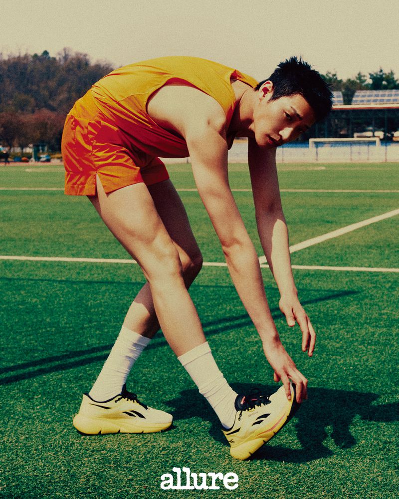 Asian man stretches outdoors on a green running track wearing yellow athletic top, orange shorts, white socks and athletic shoes.