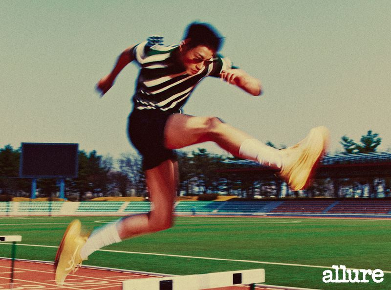 Athlete hurdles a track obstacle, wearing retro striped athletic wear, in a medium shot with daytime outdoor setting and bright lighting.