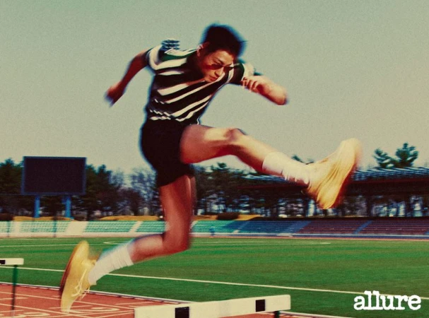 Athlete hurdles a track obstacle, wearing retro striped athletic wear, in a medium shot with daytime outdoor setting and bright lighting.