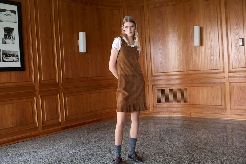 Bodine Van Galen wearing a brown suede dress over a white top with grey socks in a wood-paneled room.