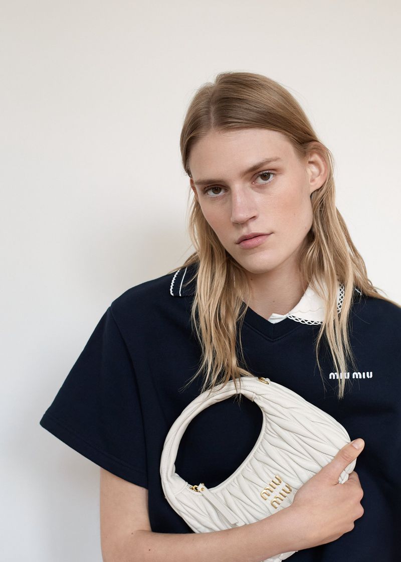 Bodine Van Galen holding a white quilted leather Miu Miu handbag while wearing a navy polo shirt against a pale studio background.