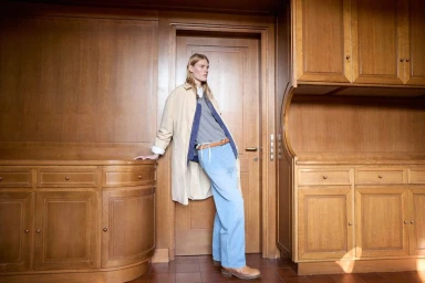 Model Bodine Van Galen leaning against a wooden doorway wearing a beige coat and light blue trousers in a wood-paneled room.