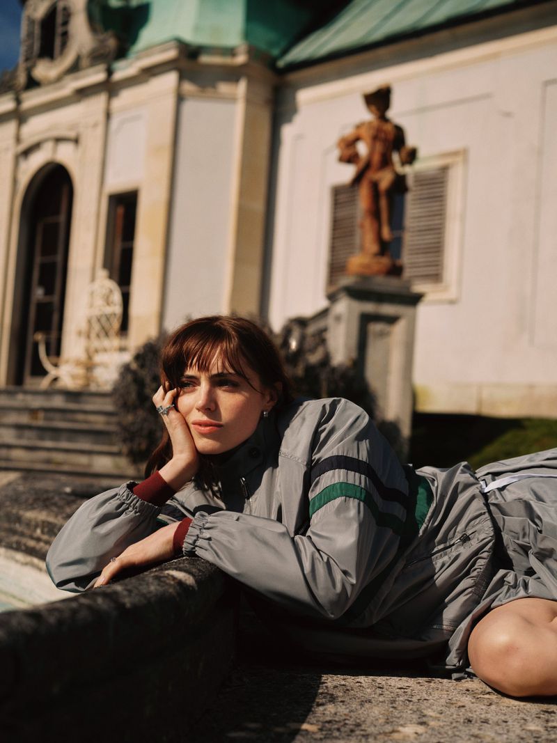 Bella Maclean wearing a grey striped track jacket resting her chin on her hand beside a stone ledge outside a grand estate.