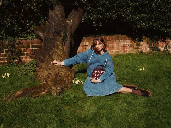 Bella Maclean wearing a blue denim quilted hoodie dress seated on a grassy lawn beside a tree trunk and brick wall.