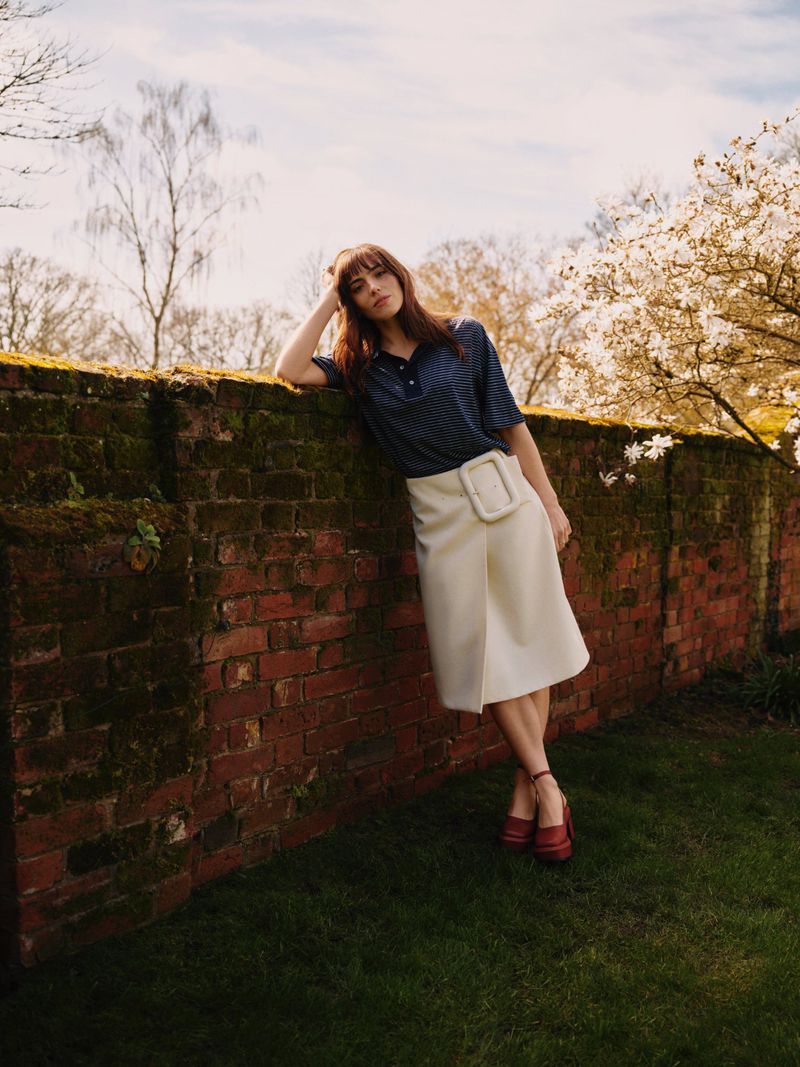 Bella Maclean leans against a mossy brick wall wearing a cream skirt with an oversized buckle and dark short-sleeve shirt.