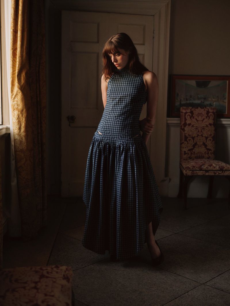 Bella Maclean standing indoors by a curtained window wearing a navy blue checked drop-waist maxi dress.
