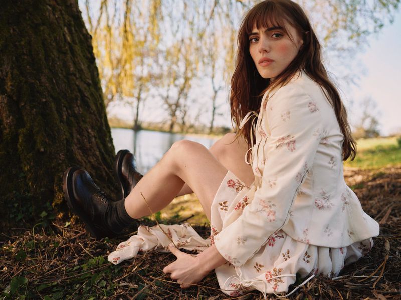 Bella Maclean seated outdoors beneath a large tree wearing a cream floral print jacket and black combat boots.