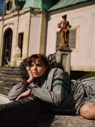 Bella Maclean wearing a grey striped track jacket resting her chin on her hand beside a stone ledge outside a grand estate.