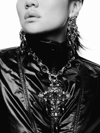 Black and white close-up of a woman wearing an ornate jeweled cross pendant necklace, matching drop earrings, and a shiny black jacket.