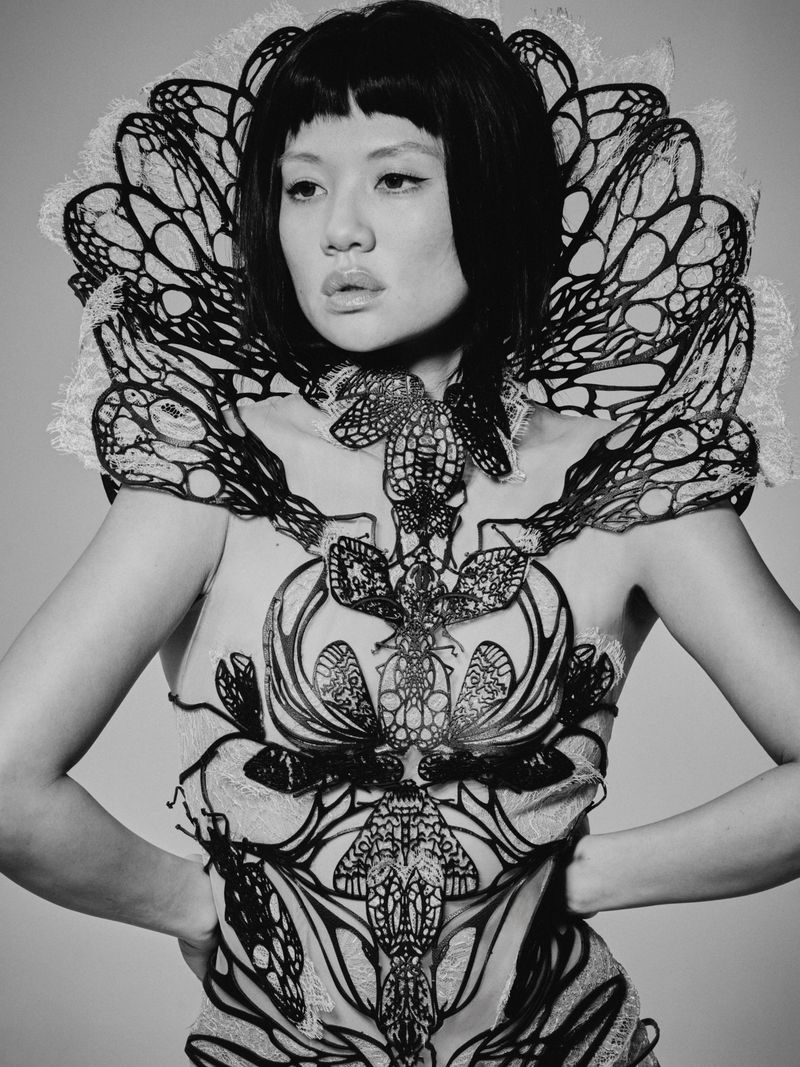 East Asian woman posing in an intricate sculptural black wing collar and corset against a grey studio backdrop.