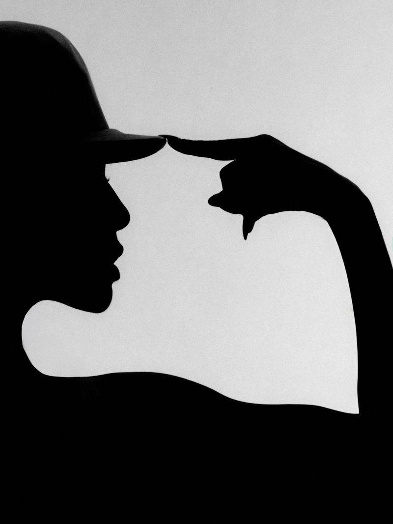 Black silhouette profile of a woman wearing a cap and touching the brim with a pointed finger against a bright seamless background.