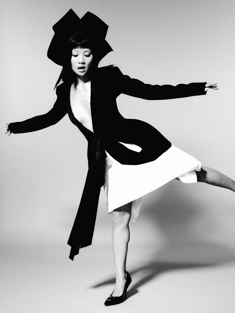 Woman in a black blazer and white skirt wearing a large geometric hat posing against a seamless grey backdrop
