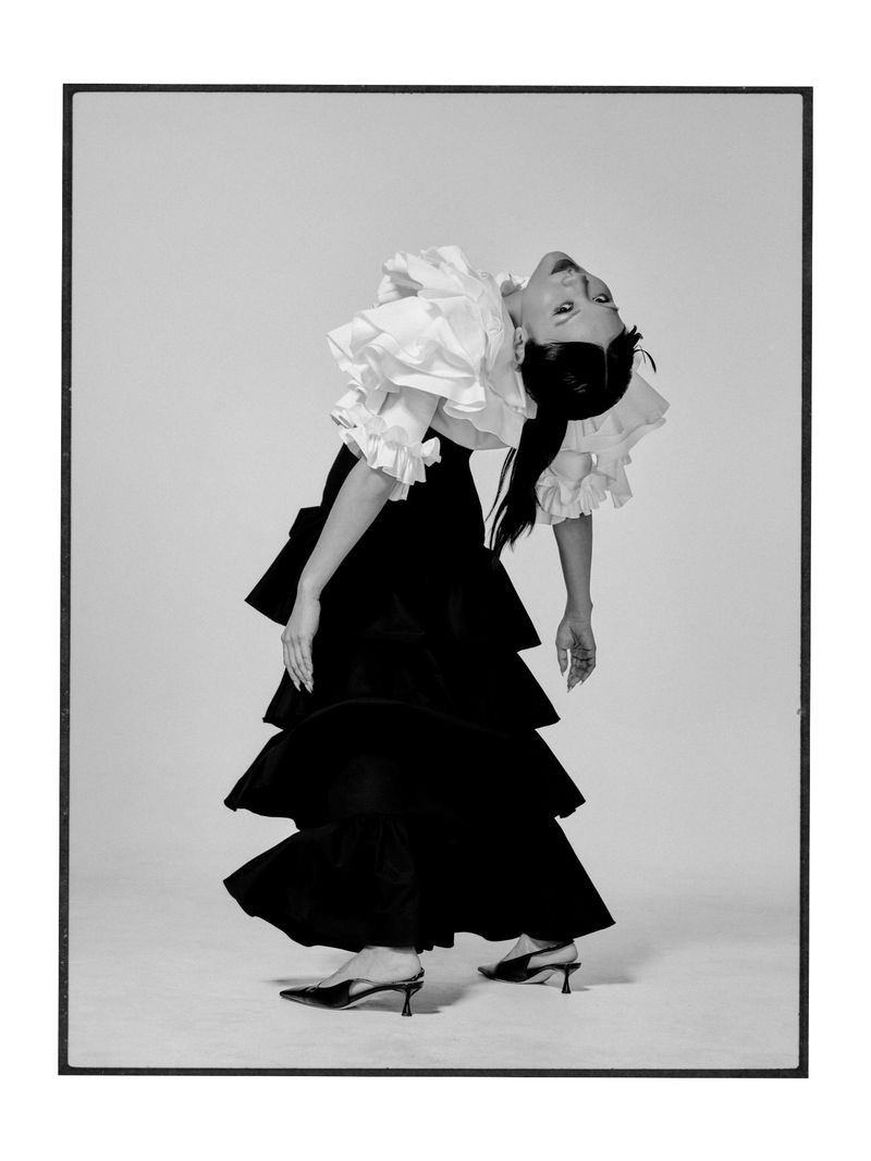 Rei Ami arching backwards wearing a white ruffled top and tiered black maxi skirt with slingback heels against a studio backdrop.
