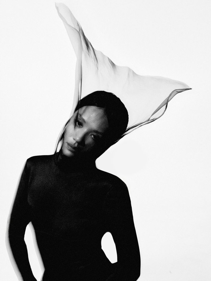 Black and white portrait of a woman wearing a dark top with a floating sheer fabric veil above her head against a white background.