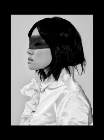 Black and white profile portrait of Rei Ami wearing a dark eye mask and a white ruffled satin blouse against a plain studio backdrop.