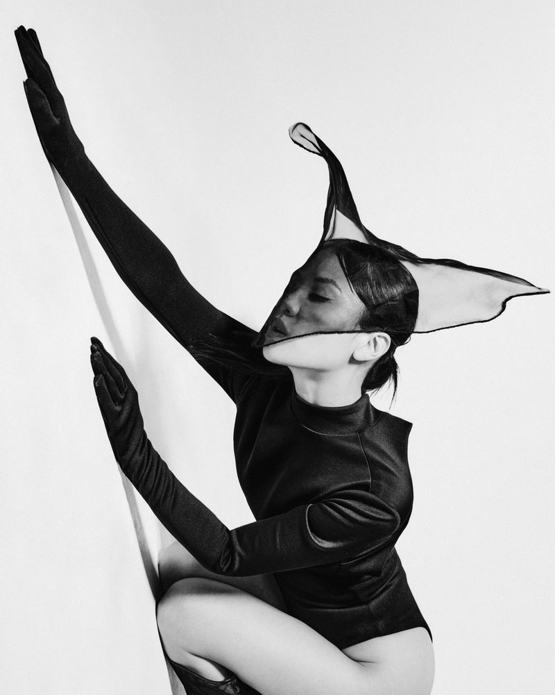 East Asian woman in a black bodysuit and gloves wearing a sheer sculptural headpiece against a white background.