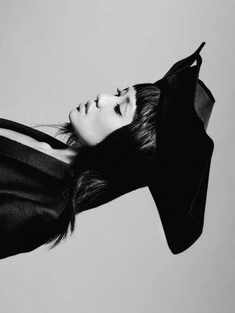 Black and white profile portrait of a woman wearing an avant-garde sculptural black hat and blazer against a seamless studio background.