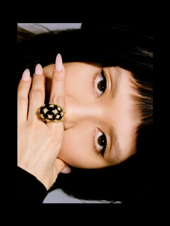 Close-up portrait of an East Asian woman with dark bangs resting her hand with a large gold and black statement ring over her face.