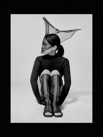 Woman seated in a black long-sleeved top and open-toe knee-high boots with an architectural mesh headpiece against a plain backdrop.
