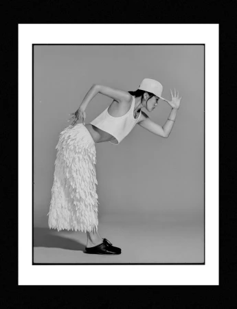Black and white fashion portrait of a woman bending forward in a cropped tank top, textured feathered skirt, and backwards cap.