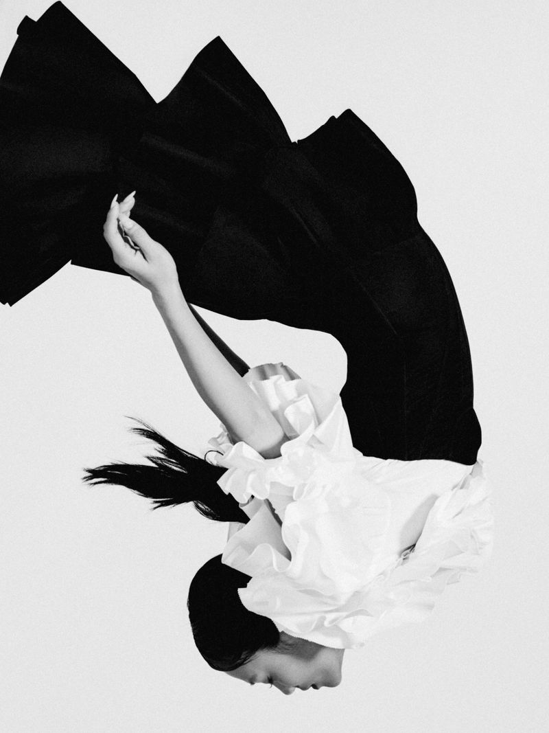 Rei Ami captured in an inverted pose wearing a ruffled white blouse and structured black dress against a plain white studio background.