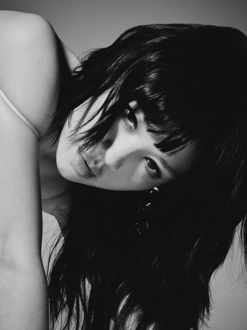 Close-up black and white portrait of a woman with dark fringe bangs leaning forward and looking directly at the camera.