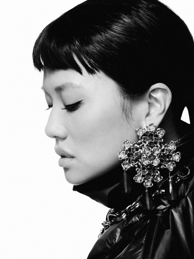 Monochrome profile portrait of REI AMI wearing an ornate jeweled cross earring and a high-collar jacket against a white background.