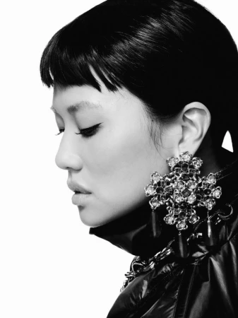 Monochrome profile portrait of REI AMI wearing an ornate jeweled cross earring and a high-collar jacket against a white background.