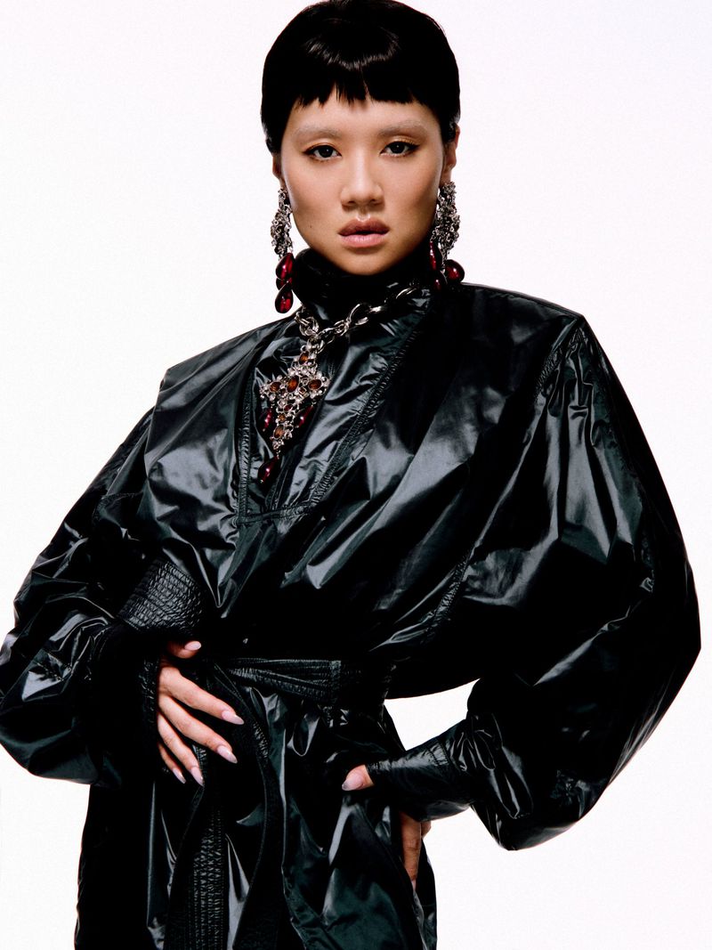 Rei Ami wearing a glossy black oversized jacket and ornate jewel necklace against a solid white studio background.
