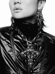 Black and white close-up of a woman wearing an ornate jeweled cross pendant necklace, matching drop earrings, and a shiny black jacket.