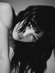 Close-up black and white portrait of a woman with dark fringe bangs leaning forward and looking directly at the camera.
