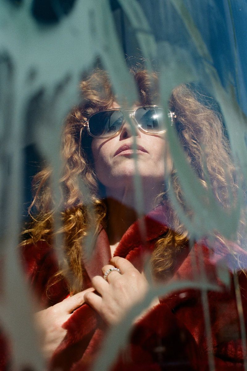 Portrait of a woman with curly hair and sunglasses, wearing a red fur coat, gazing upward with light shining and reflection.