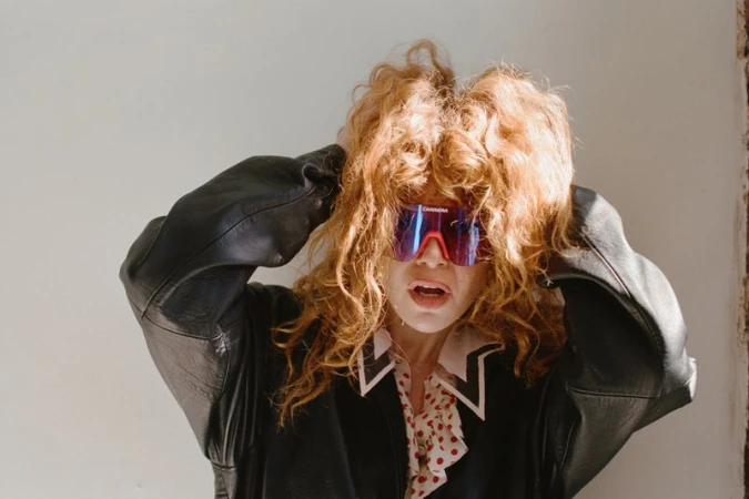 Natasha Lyonne wearing reflective visor sunglasses and a black leather jacket with hands in her curly reddish hair.