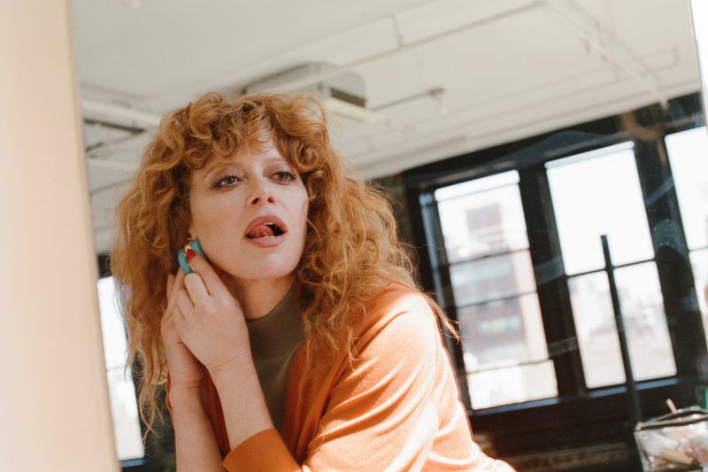 Natasha Lyonne adjusting a blue hoop earring while looking in a mirror wearing an orange cardigan.