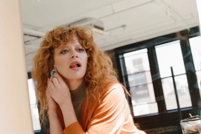 Natasha Lyonne adjusting a blue hoop earring while looking in a mirror wearing an orange cardigan.