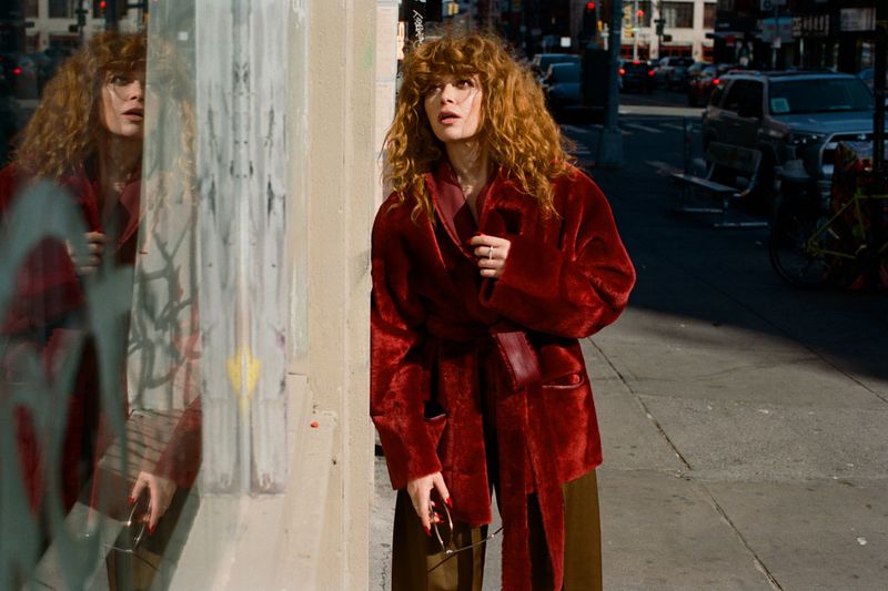 Natasha Lyonne wearing a belted red coat looking into a storefront window on a sunlit city street.