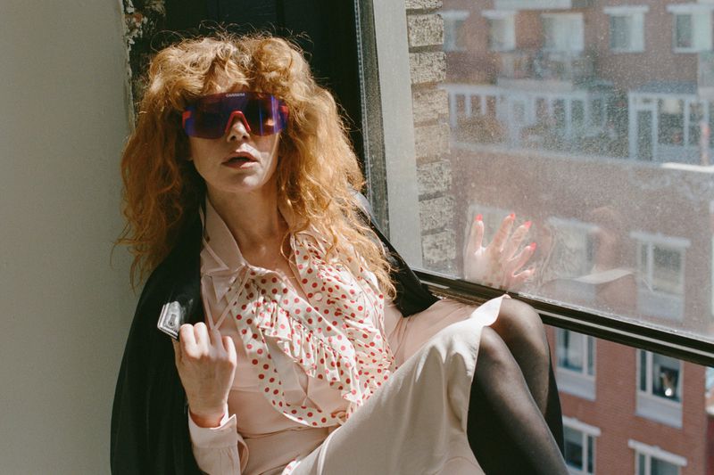 Natasha Lyonne wearing sports sunglasses and a polka dot dress while seated indoors by a large window.