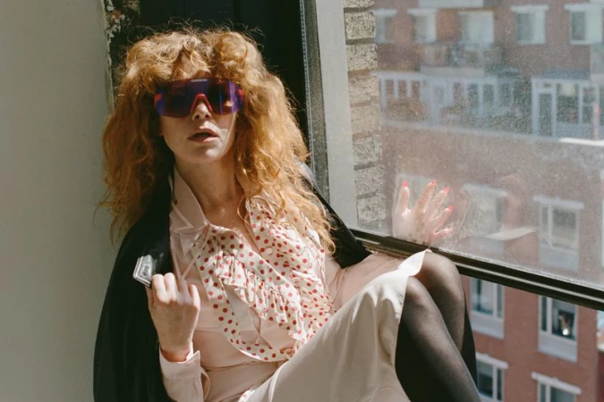 Natasha Lyonne wearing sports sunglasses and a polka dot dress while seated indoors by a large window.