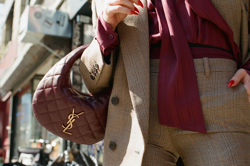 Natasha Lyonne wearing a brown plaid suit and burgundy bow blouse while holding a quilted burgundy leather Saint Laurent handbag.
