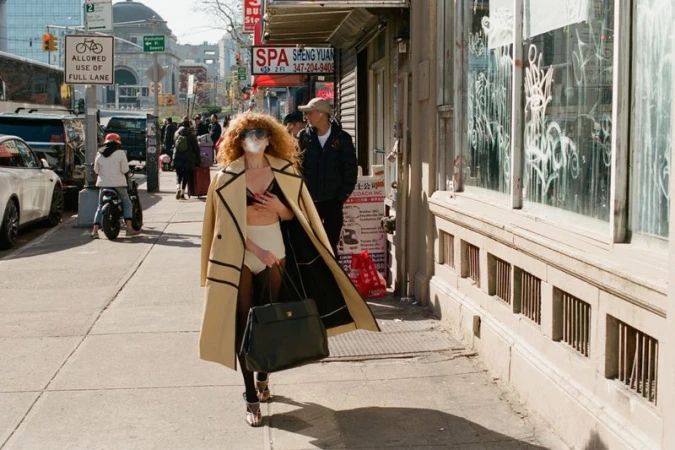 Natasha Lyonne walking down a New York city sidewalk wearing an unbuttoned beige coat and black handbag