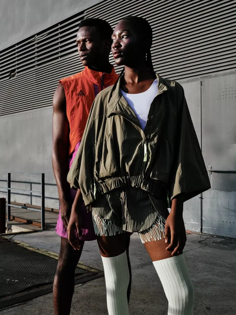 A man and woman posing outdoors against a slatted metallic exterior wall, wearing sportswear and high socks