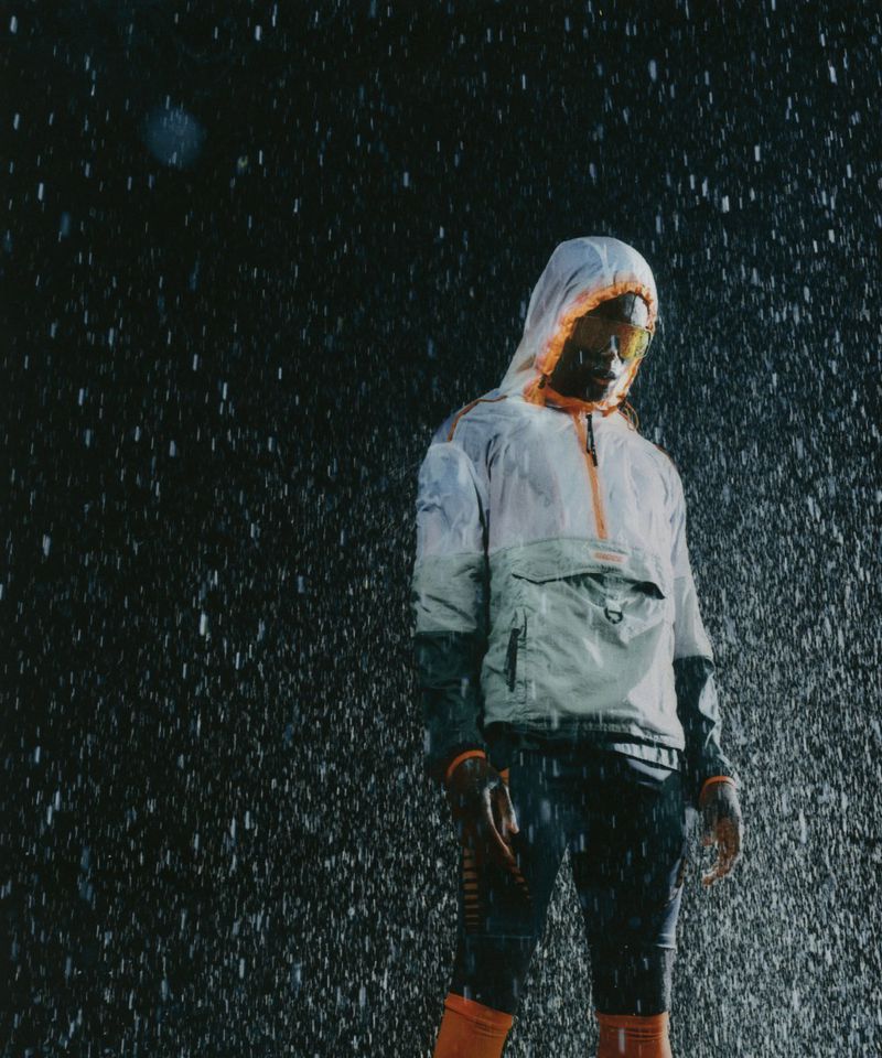 A man wearing a hooded technical rain jacket and tinted visor standing in heavy falling rain against a dark background.