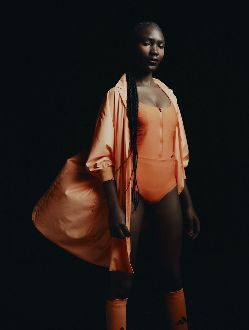Black woman wearing an orange one-piece swimsuit and matching lightweight hooded parka against a solid black background.