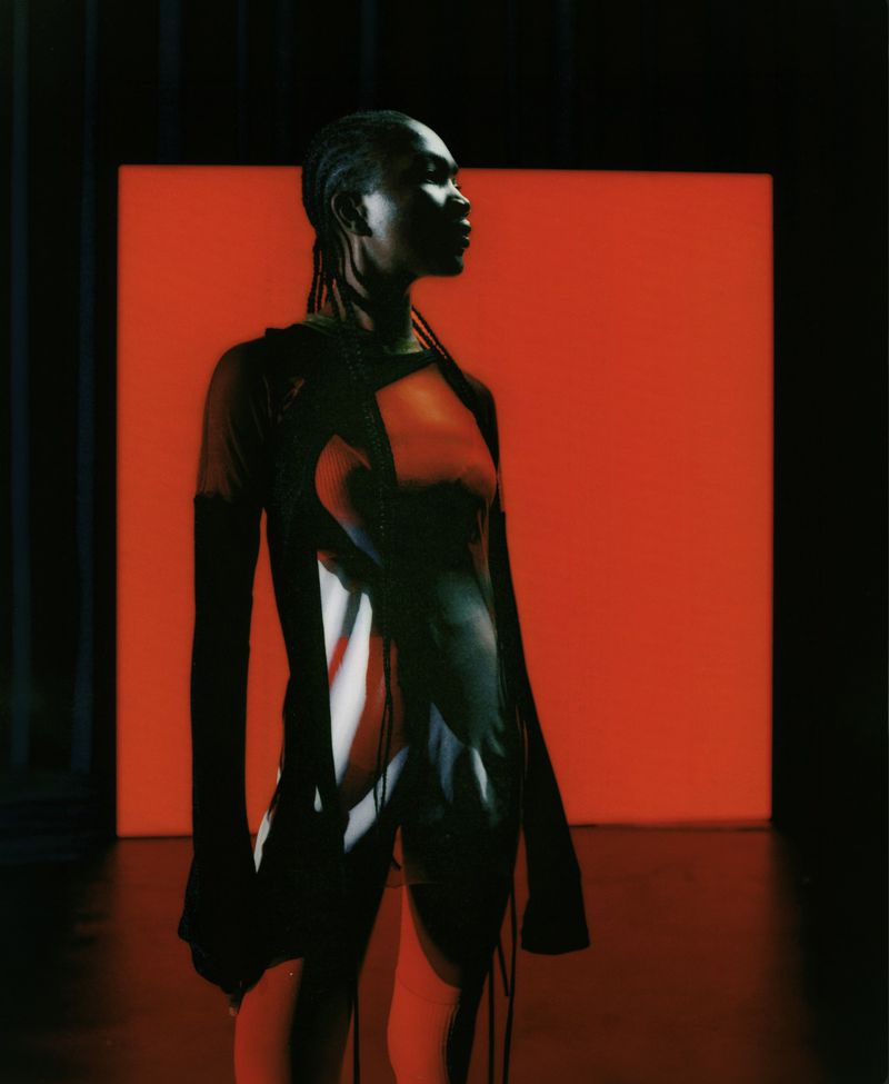 A Black model in a graphic printed dress and black arm warmers poses in profile before a vivid red glowing square in a dark studio.