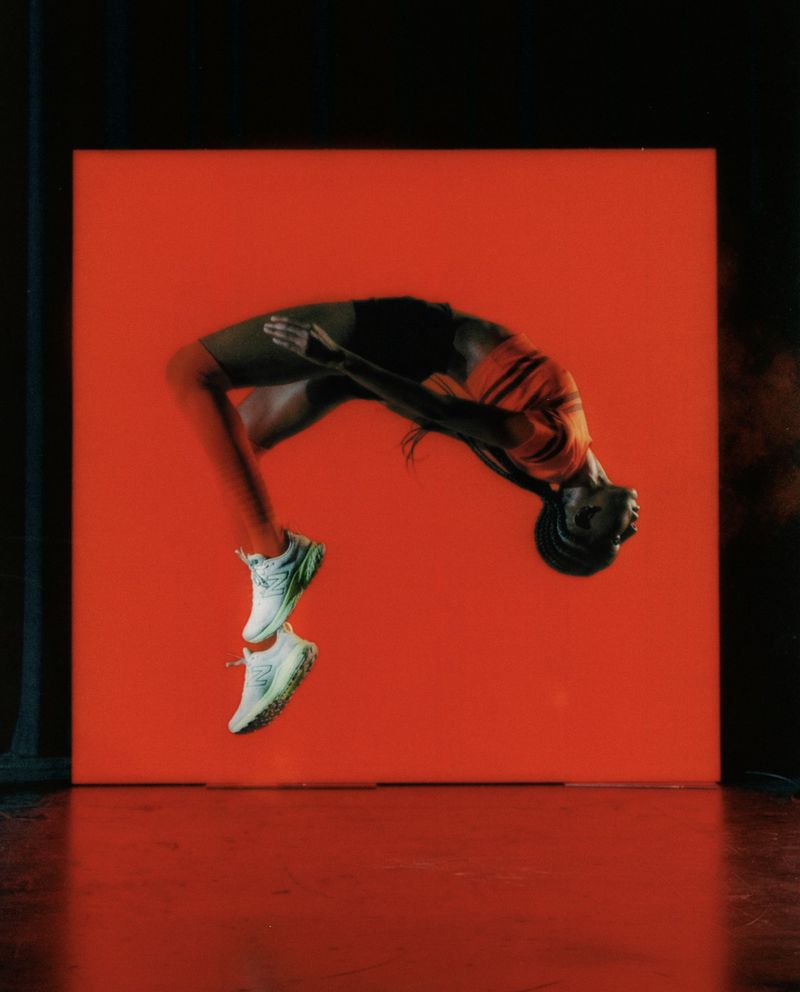 An athlete suspended mid-air in an arched jump wearing white running shoes against a vibrant glowing orange-red square panel.