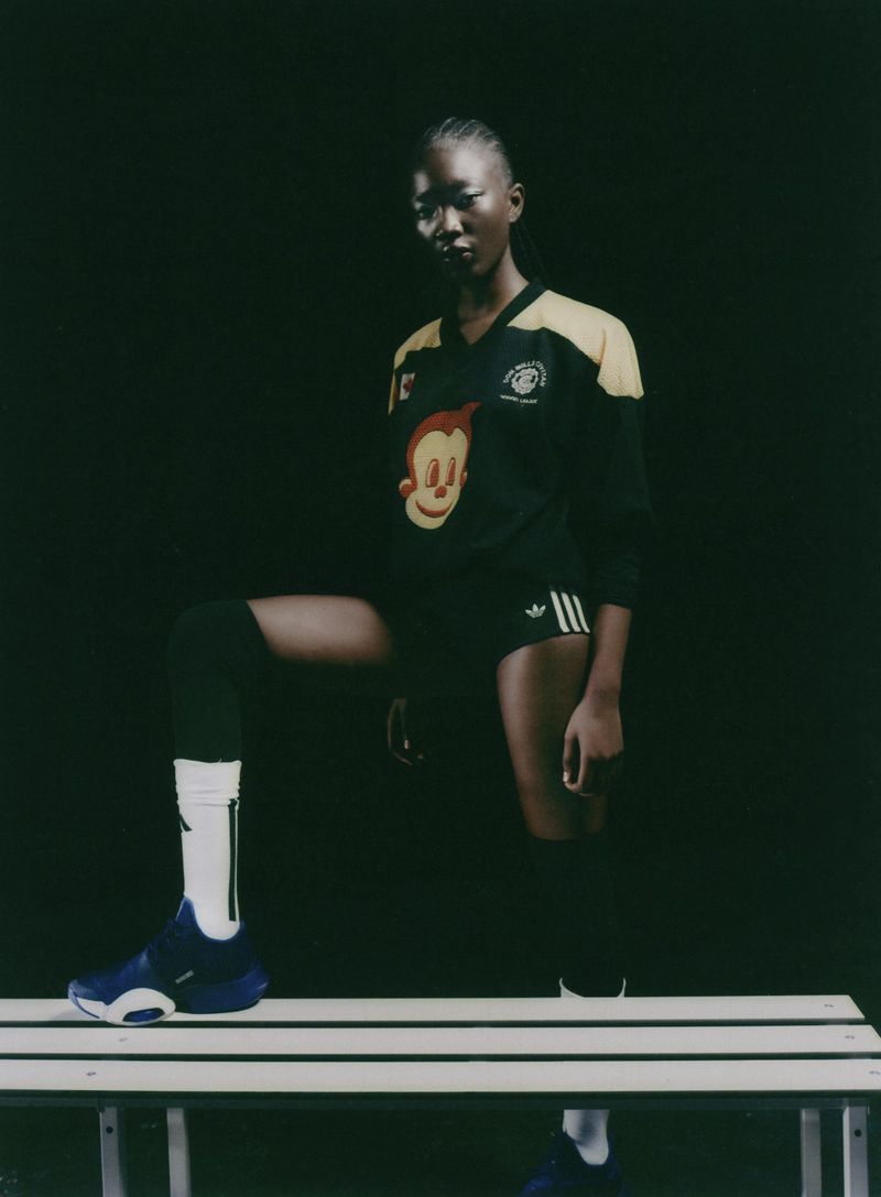 Model posing with one foot on a white bench wearing a black athletic jersey and shorts against a dark background.