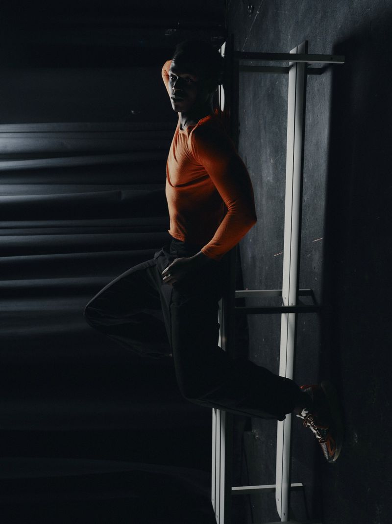 A Black man wearing an orange long-sleeve athletic shirt and dark trousers sits on a bench in a low-key, dark setting.