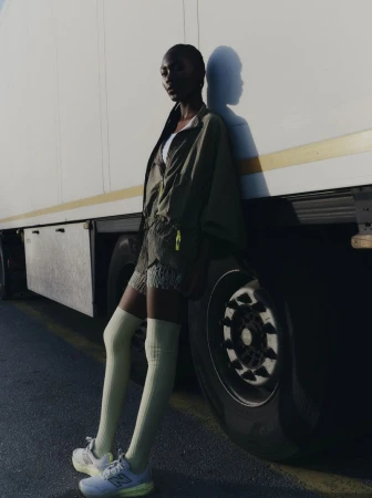 A Black woman in an olive jacket, pale green knee-high socks, and sneakers leans against the side of a large white truck in direct sunlight.